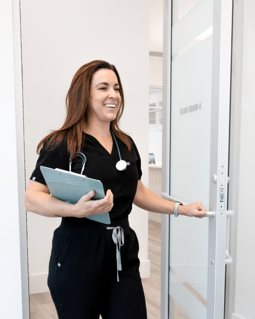 2 - Sweetgrass Plastic Surgery Smiling female nurse in black scrubs holds a clipboard and opens an exam room door labeled 'EXAM ROOM 4'.