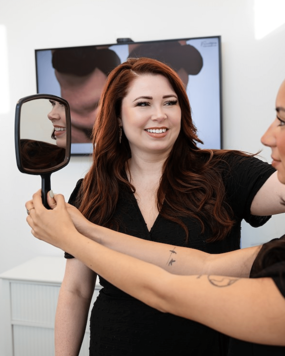 - Sweetgrass Plastic Surgery Woman in black smiles as a stylist holds a handheld mirror to her left, showing a friendly salon moment.
