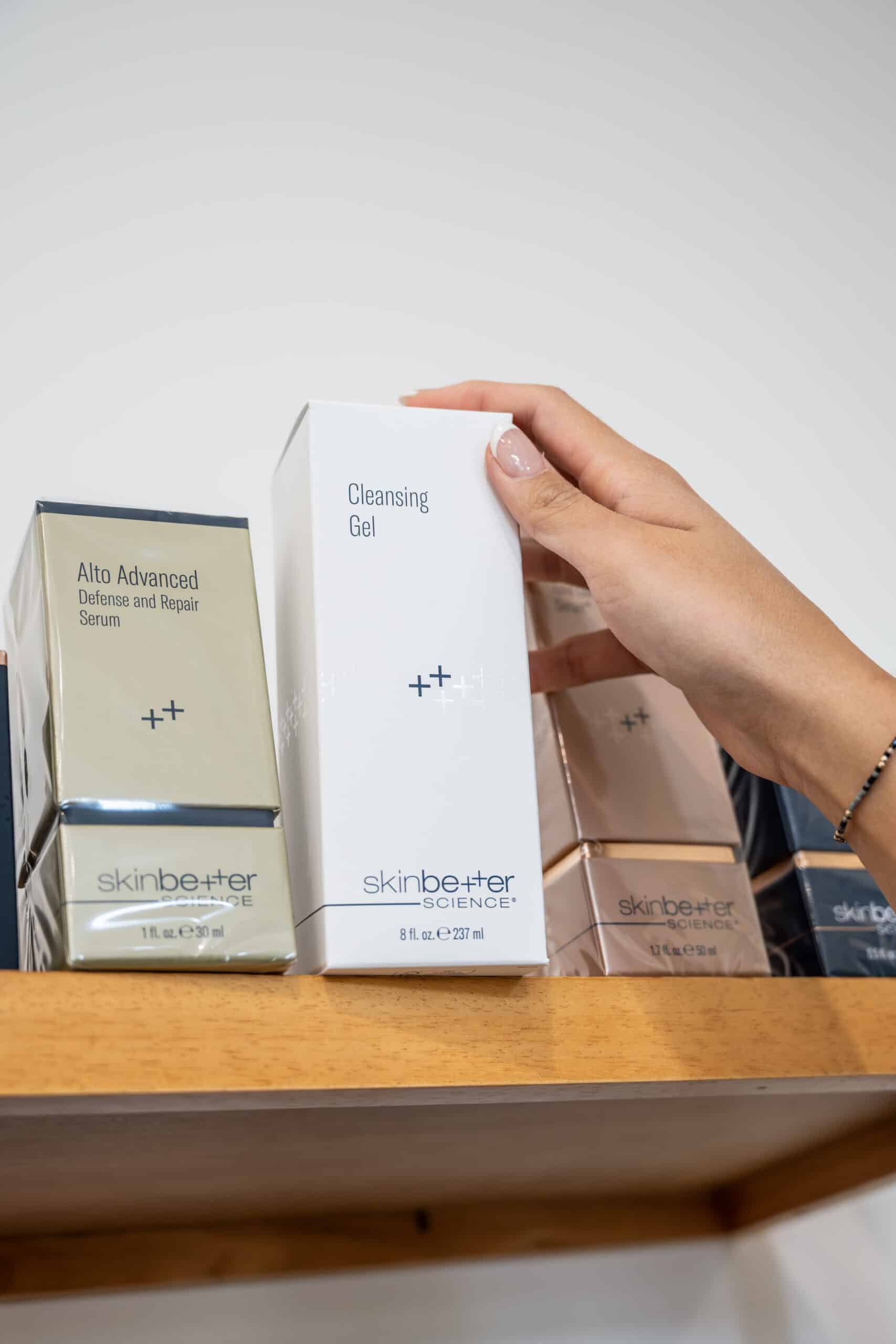 Skincare - Sweetgrass Plastic Surgery Hand holding a white box labeled Cleansing Gel among skincare product boxes on a shelf.